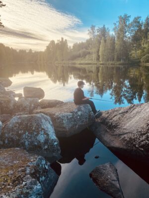 Poika istuu järven rannan kivikoilla ja kalastaa